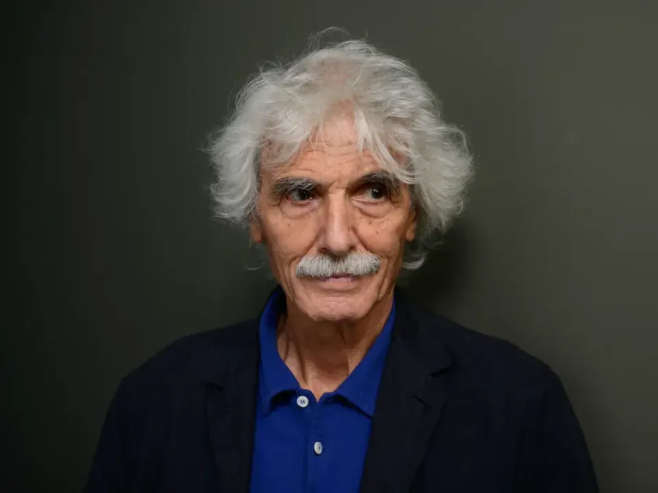 A smiling man with long gray hair and a thick gray mustache, wearing a blue collared shirt with a dark jacket over it, standing against a neutral backdrop..