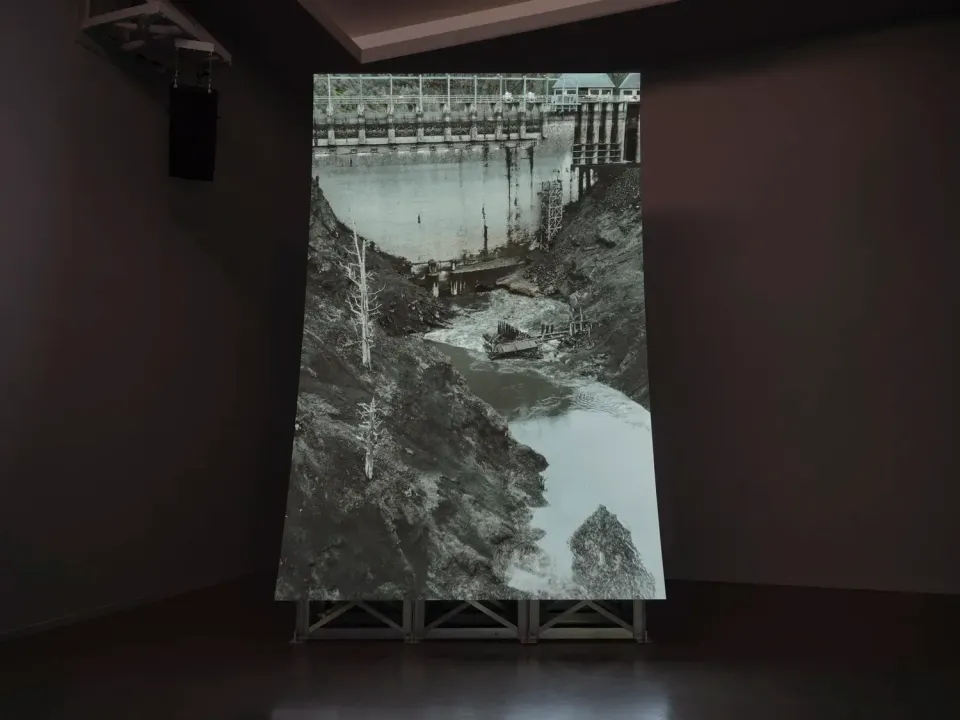 A vertical screen installed in a dark gallery space, with a black-and-white image of a river and dam.