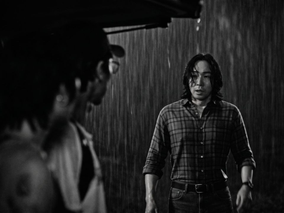 A black and white image of a man with long hair wearing a plaid shirt and standing in the rain.