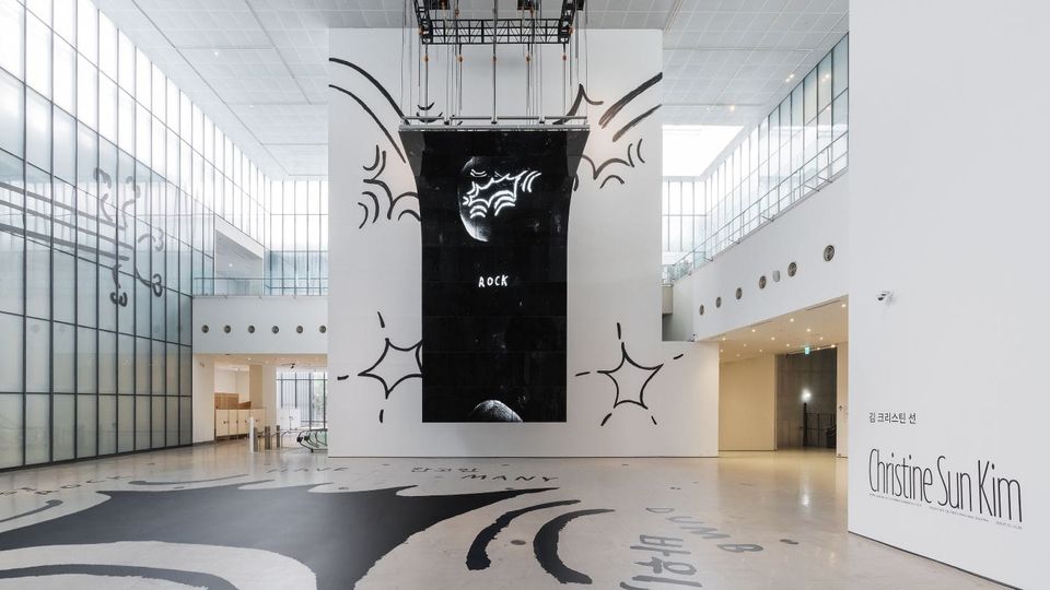 View of a large art gallery with illustrations on the floor and wall and a large black banner with the word "ROCK" hanging from the ceiling.