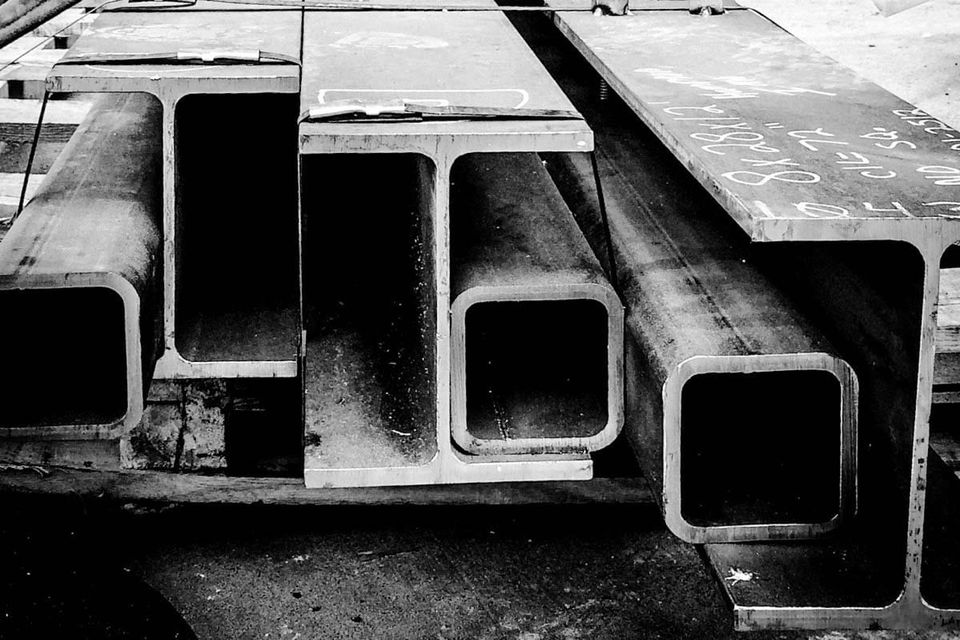 Black and white close-up photograph of stacked structural steel I-beams and hollow rectangular tubing, shot from a low angle."