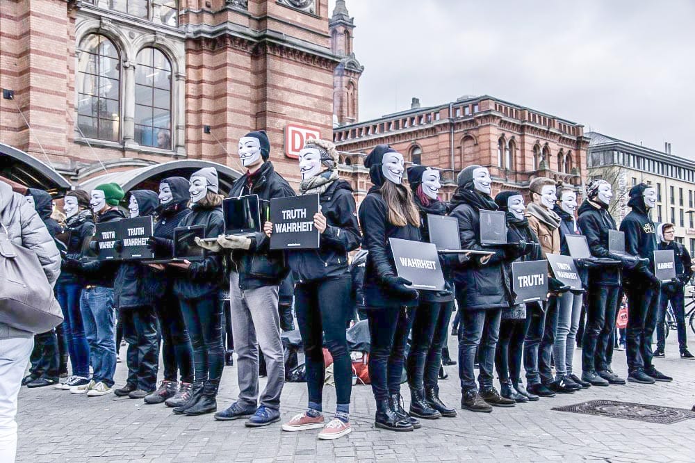 People in black clothes and with Guy Fawkes masks holding up signs and monitors. The signs read "TRUTH | WAHRHEIT".