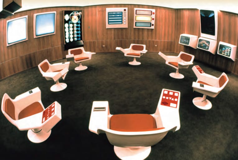 The retro futuristic hexagonal Operations Room of Project Cybersyn. 7 turnable chairs in a circle, monitors and control elements on the walls. A room designed to hold complexity under load.