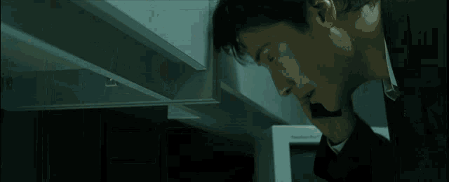 Neo at his desk in the tie, the boss's shadow arriving. The agents turn. He hides.