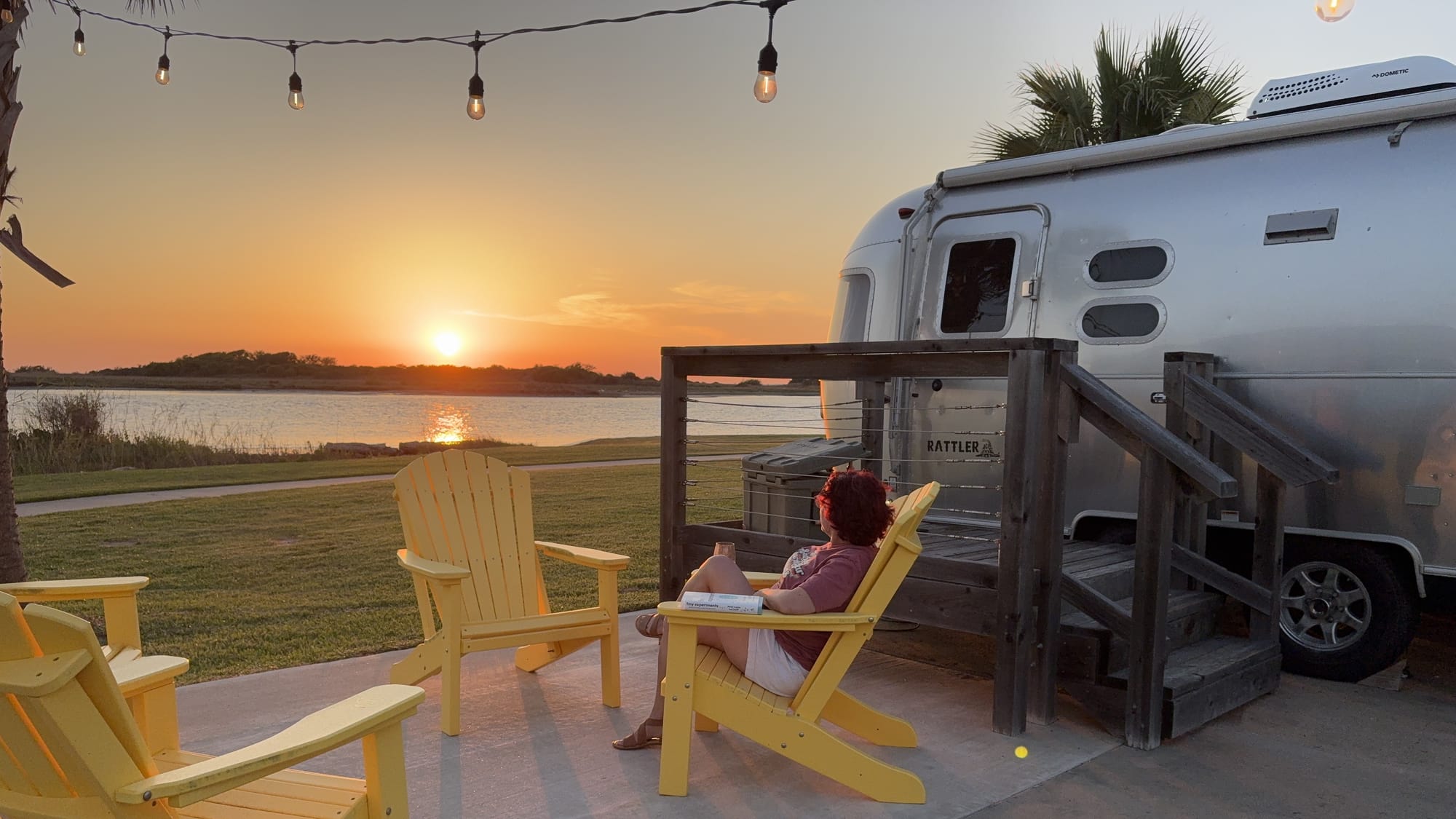Woman sitting in yellow Adirondack chair outside a silver Airstream travel trailer watching sunset.