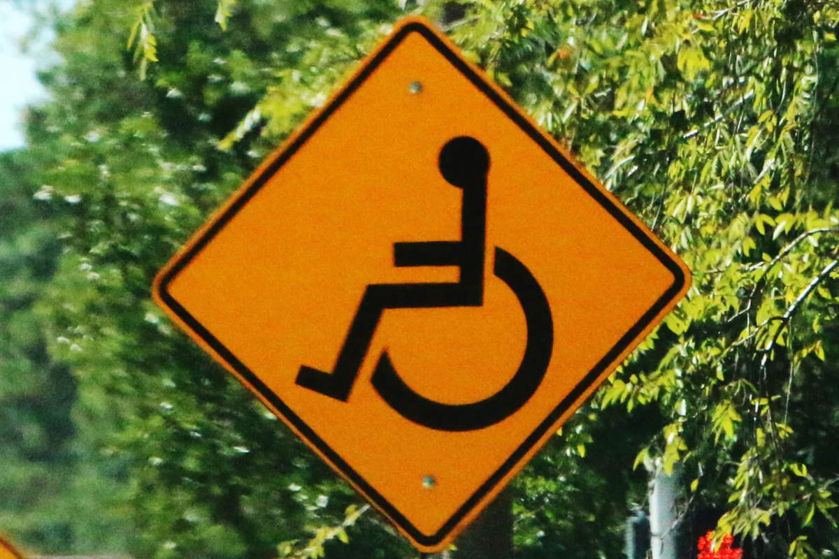Black wheelchair symbol on orange diamond sign with green bushes in background