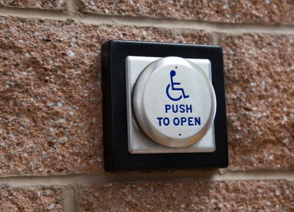Round metal Push To Open button with blue lettering and wheelchair symbol, on a brick wall