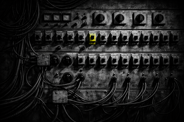 Black-and-white industrial control panel with rows of switches and cables; one central switch stands out in vivid acid yellow under harsh side lighting