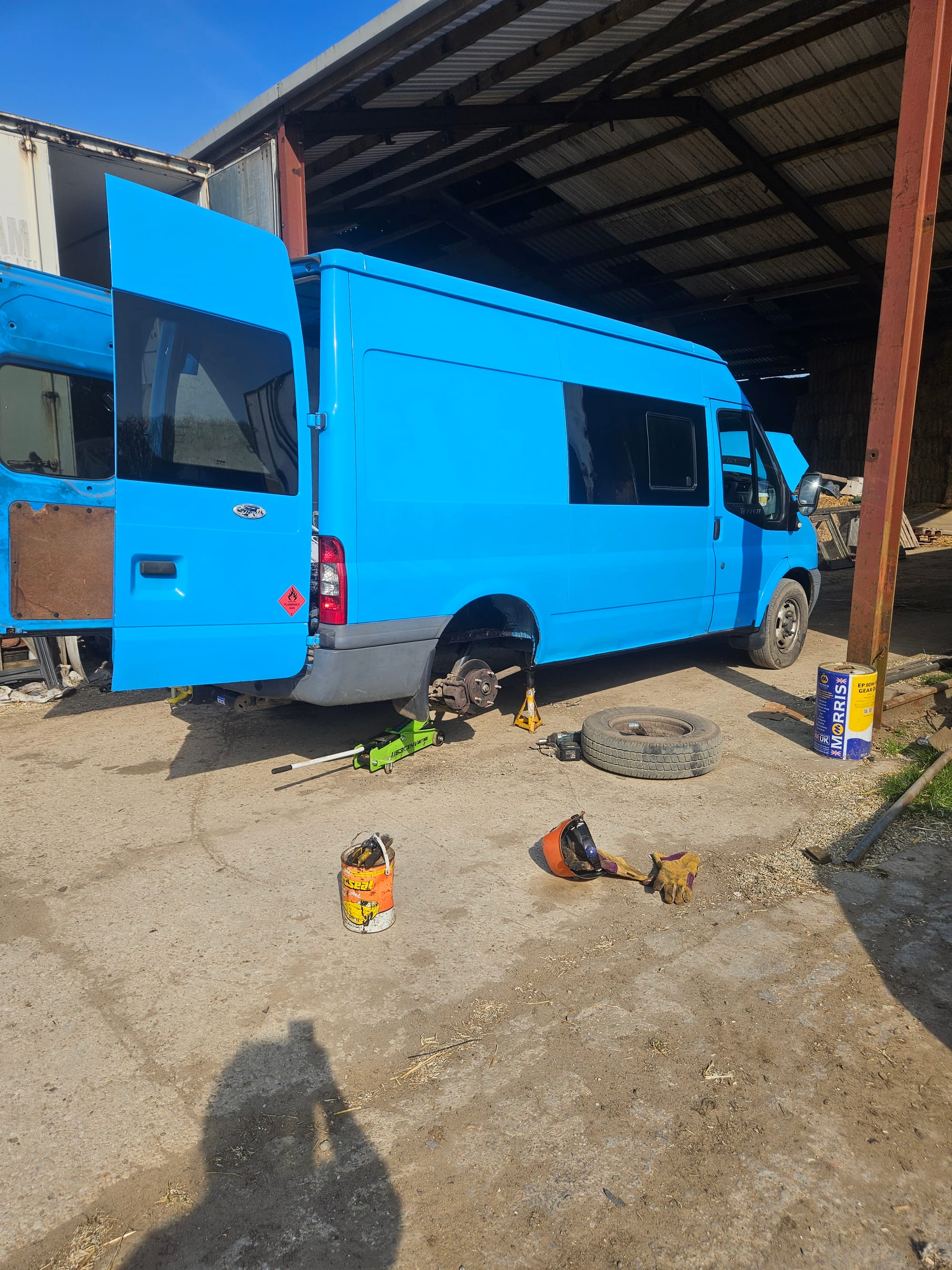 A blue panel van sits on axle stands with wheels removed and bonnet open