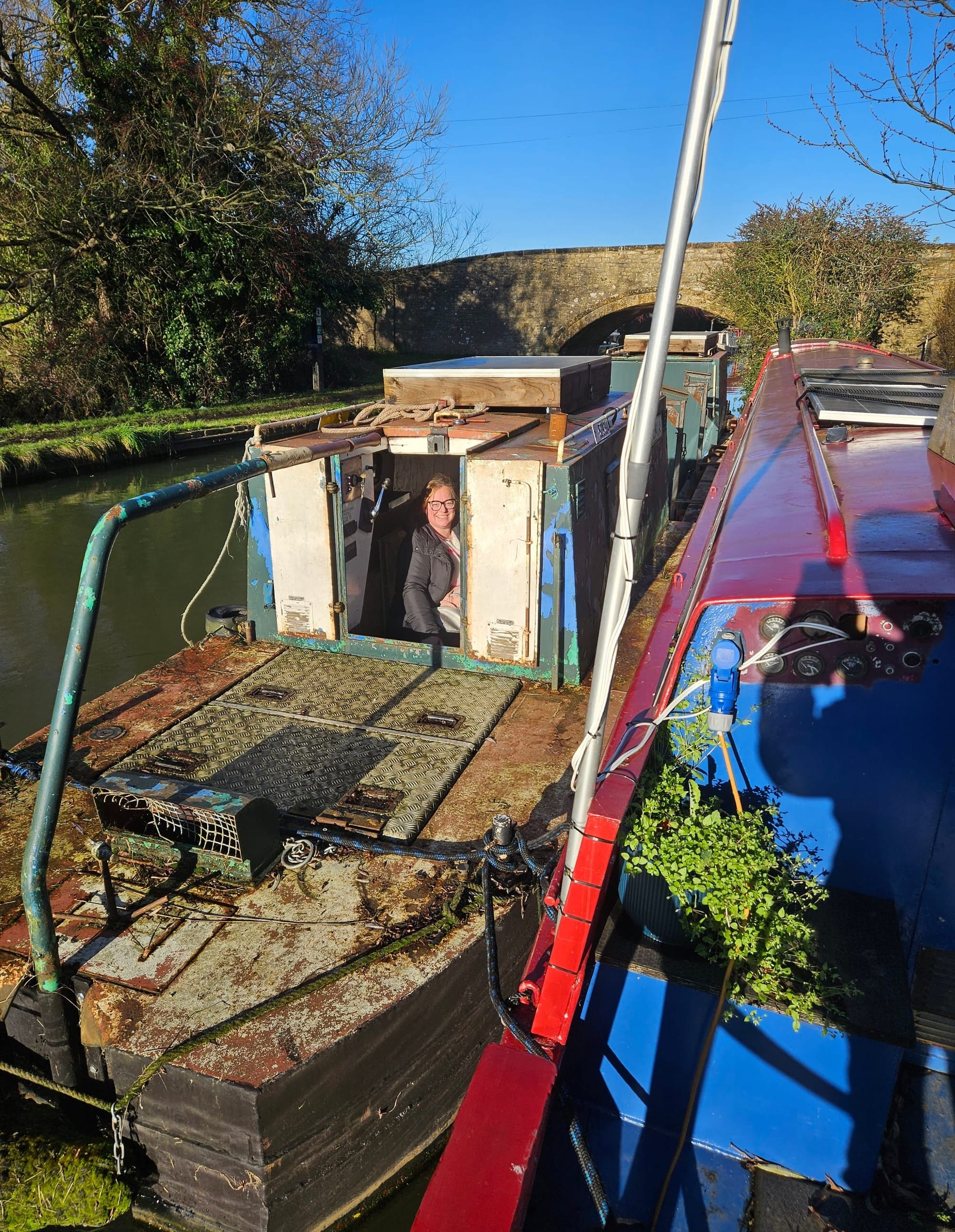 A rural scene on a canal, with a red boat tied to a green and red boat. The sky is clear and bright, and there is a stone arched bridge immediately behind the boats 