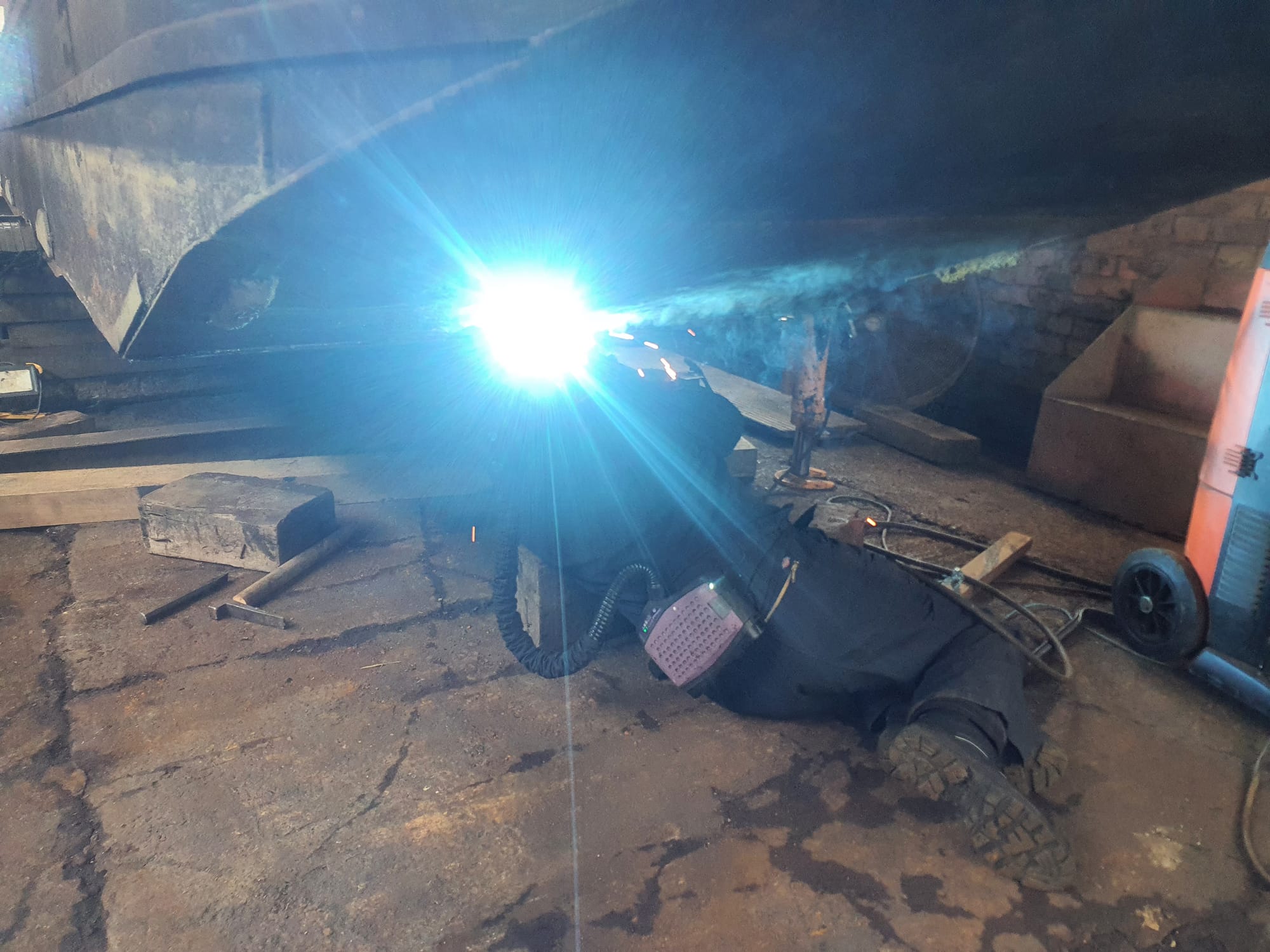 A man lies underneath a metal boat hull. He is welding the boat, and the bright arc of the welder is visible