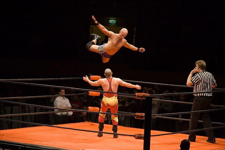 A wrestler in just shorts and kneepads is suspended in midair, while another man in a red and yellow singlet prepares for him to fall on top of him. A referee waits in another corner. 