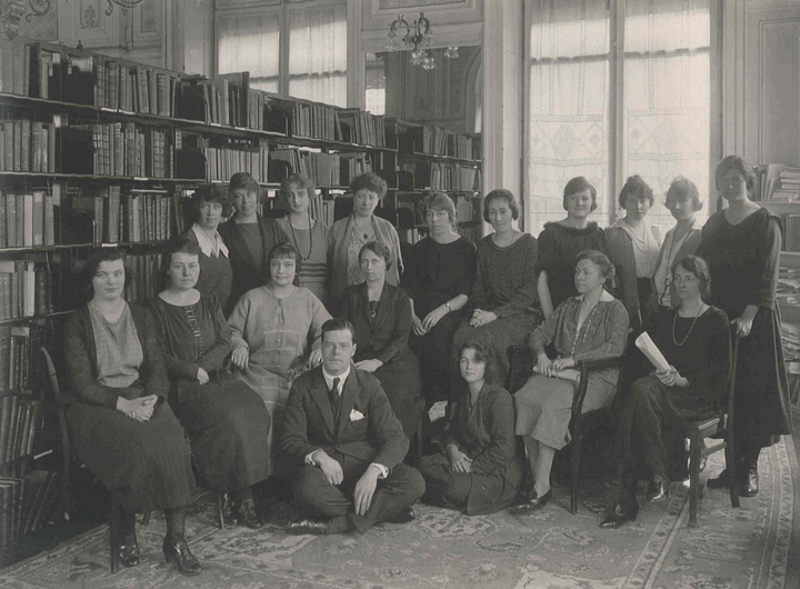 A black and white photo of 18 people seated in a group in a library, circa 1920.