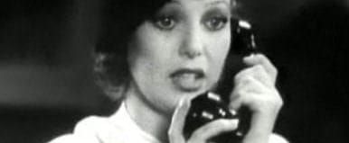 A white woman in a white blouse and black hat holding an old telephone receiver to her ear. A still from a black & white movie from the 1930s.