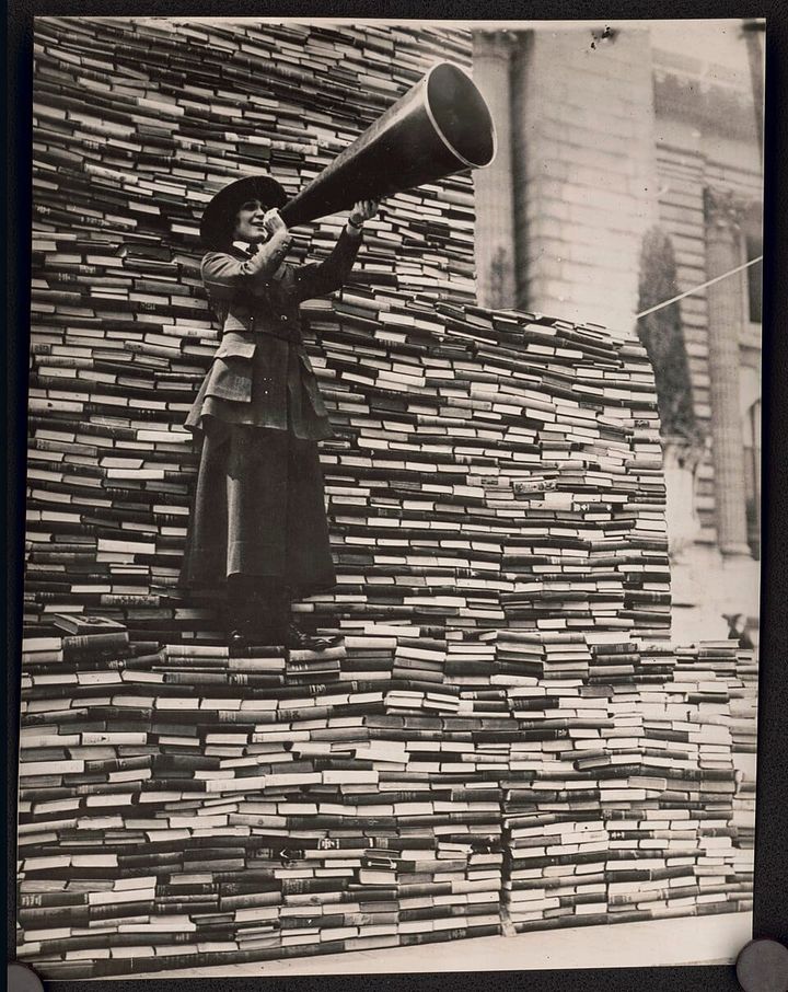 I woman in a long black coat and skirt holding a megaphone standing on a huge pile of books. Black & White.
