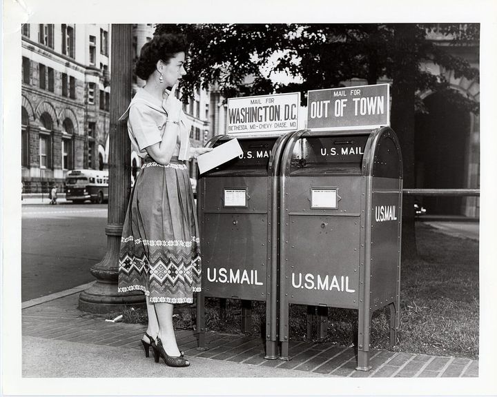 A black and white photo of a white woman in a long skirt, holding a letter, deciding which mailbox to put it in. The mailboxes are labeled Washington, DC and Out of Town, and U.S. Mail.