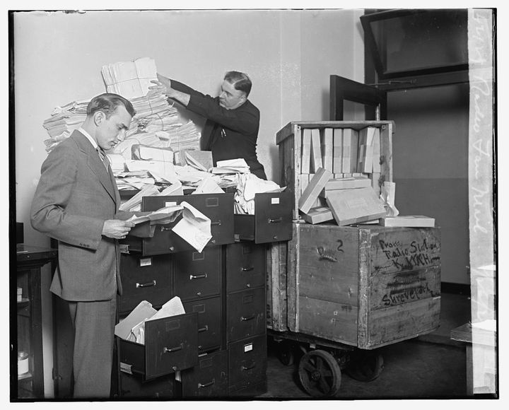 Two white men in suits standing by several file cabinets overflowing with letters and papers. 