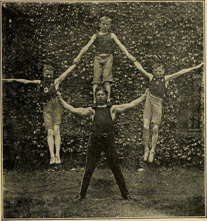 A black and white picture of a male gymnast holding one boy on his shoulders and one in each hand.