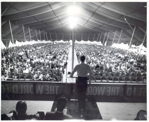 A black and white photo of a man at a podium talking to a large crowd of people in a tent.