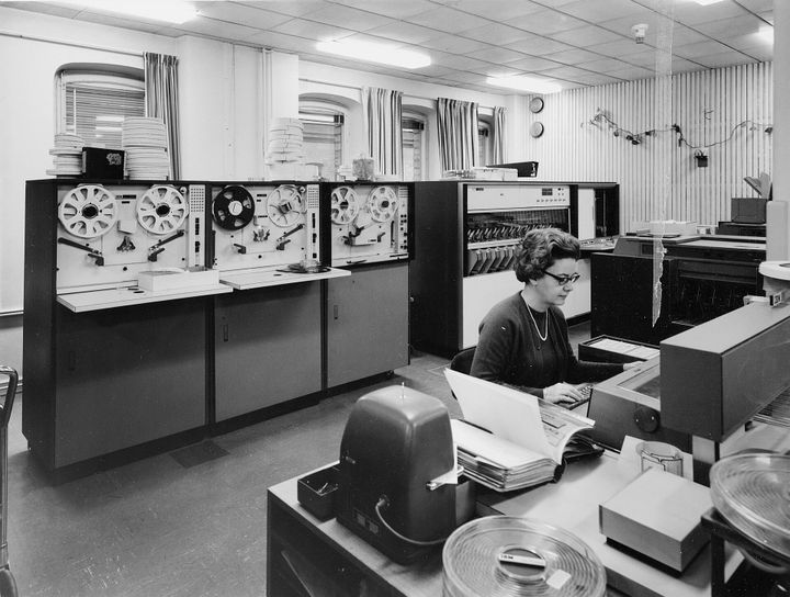 A black and white photo of a large IBM computer from the 1960s with a white woman sitting at desk at a typewriter in front of it. 