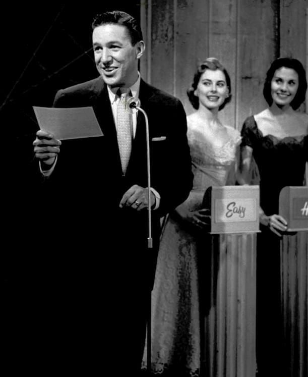 A black and white photo of a man holding a cue card and a skinny microphone. He is a game show host and there are two white women behind him holding cards that say Easy and Hard. 