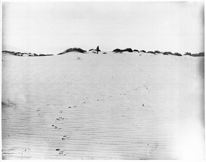 Footprints in the sand leading to a person in a hat walking over a sand dune. 