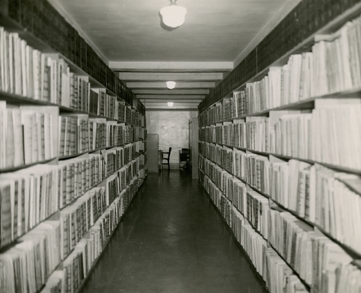 A black and white photo of a long hallway of bookshelves, ending in a desk and chair.