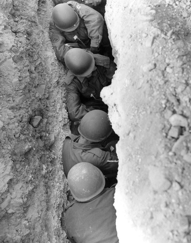 U.S. Army troops in a trench at the Nevada Test Site during Operation Teapot, 1955, rehearsing for an atomic detonation at close range — the permissible man, made literal. U.S. National Nuclear Security Administration / Nevada Site Office; source (public domain).