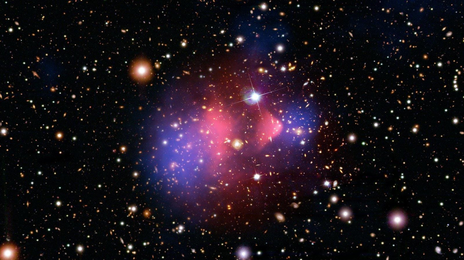 The Bullet Cluster (1E 0657-56). Pink: the hot gas seen in X-rays, which collided and lagged behind; blue: the mass reconstructed from gravitational lensing, which passed through. Both colors are assignments, not appearances. Credit: X-ray — NASA/CXC/CfA/M. Markevitch et al.; lensing map — NASA/STScI, ESO WFI, Magellan/U. Arizona/D. Clowe et al.