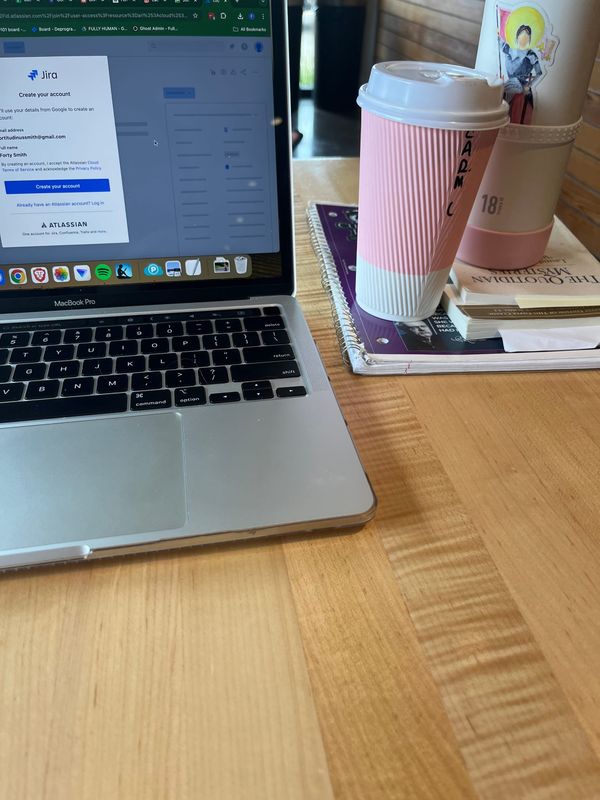 Laptop open to organization software. Stack of notebook, books, disposable pink and white coffee cup, and pink and white water bottle with watercolor Joan of Arc sticker.