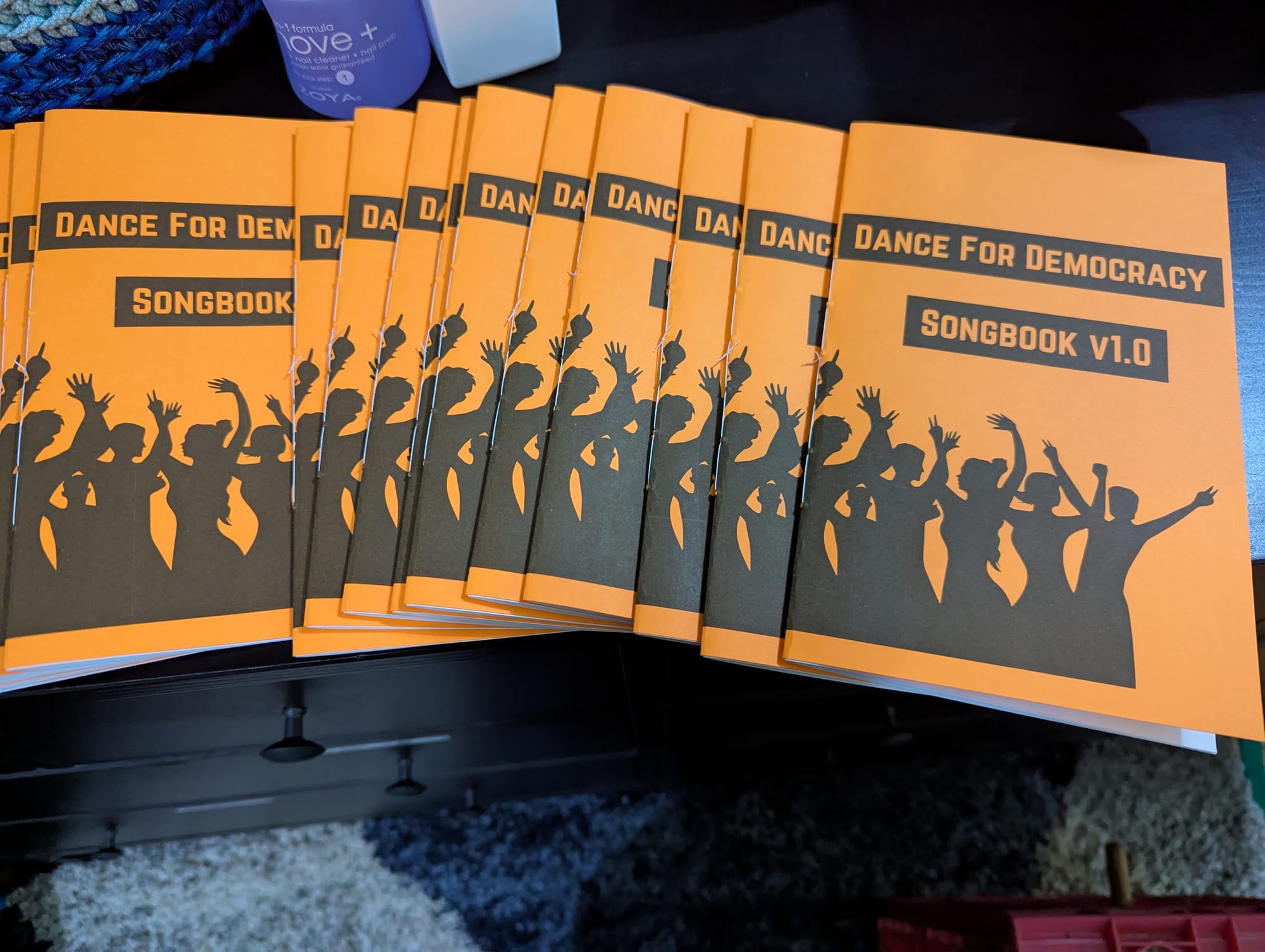 Several dozen copies of the "Dance For Democracy Songbook", a hand-bound zine with an orange cover showing the silhouettes of dancing people