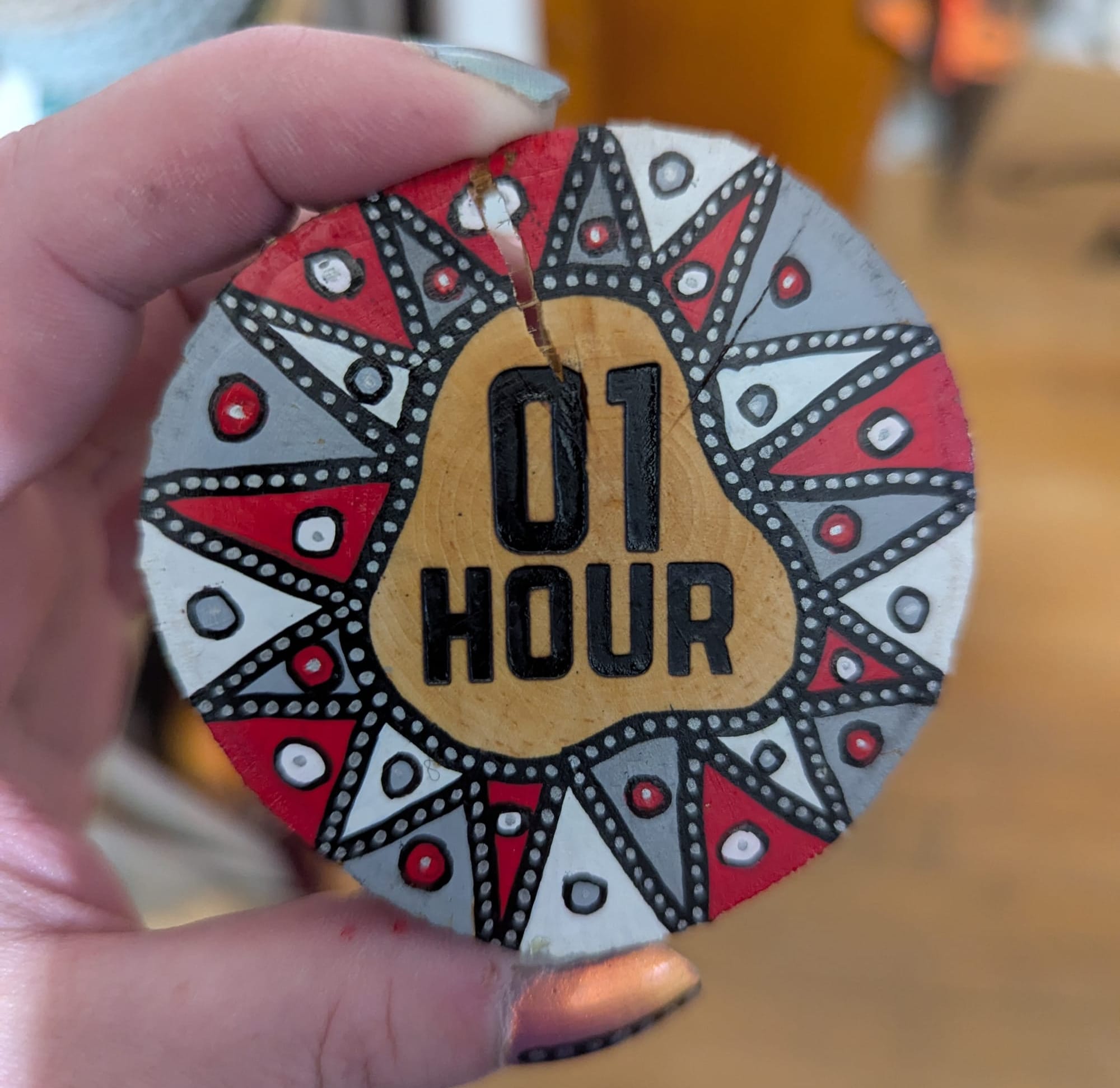 Wiley's hand holding a painted slice of serviceberry wood. At the center is a woodburned stamp reading "01 Hour". Around the edge is an elaborately painted border of red, grey, and white triangles 