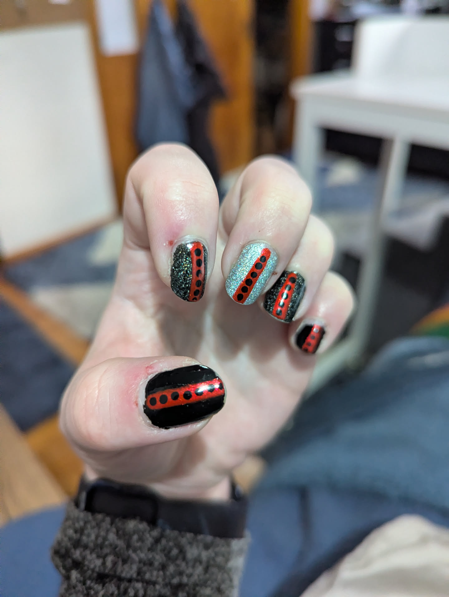 A picture of Wiley's nails, with black, holographic charcoal, and holographic silver polish. Each nail features a red central stripe with black dots on it.