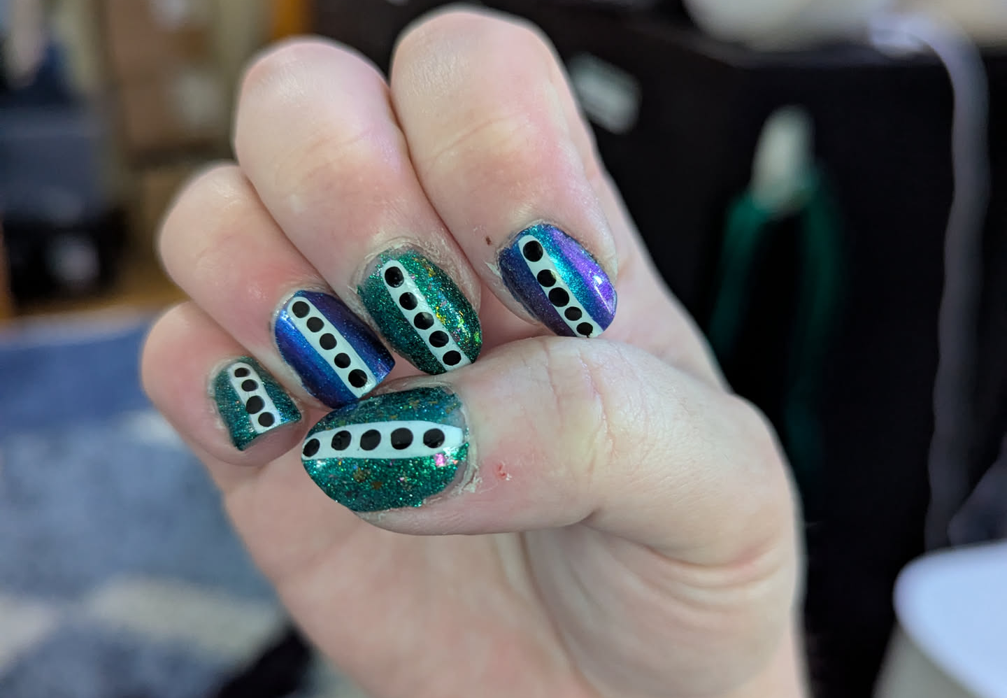 A hand with elaborately painted fingernails. The odd-numbered fingers are sparkly green, while the even numbered fingers are sparkly blue. They all have a white stripe down the middle with black dots on it.