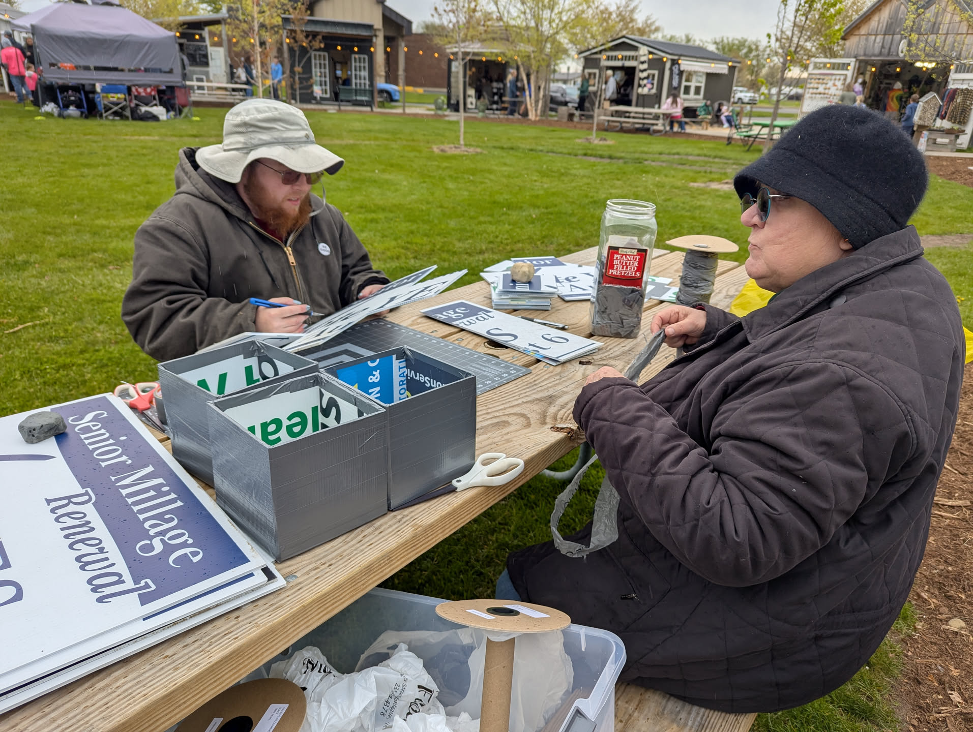 Two people sitting around a picnic table. One is turning plastic yard signs into boxes, and the other is turning plastic bags into yarn.