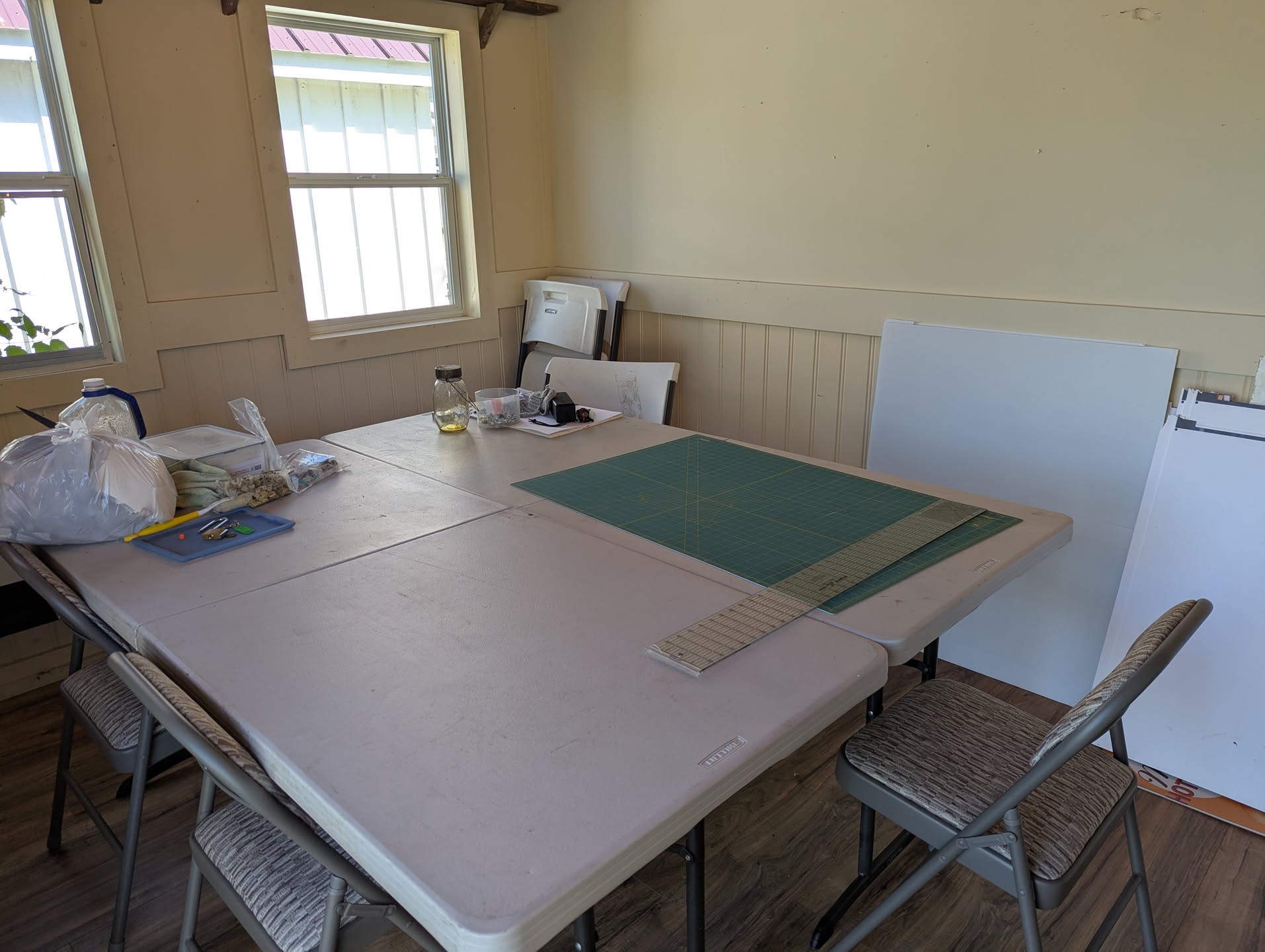 Two folding tables have been pushed together to form a big square table. Around it are folding chairs. There's a cutting mat and a quilting ruler on one side of the table.