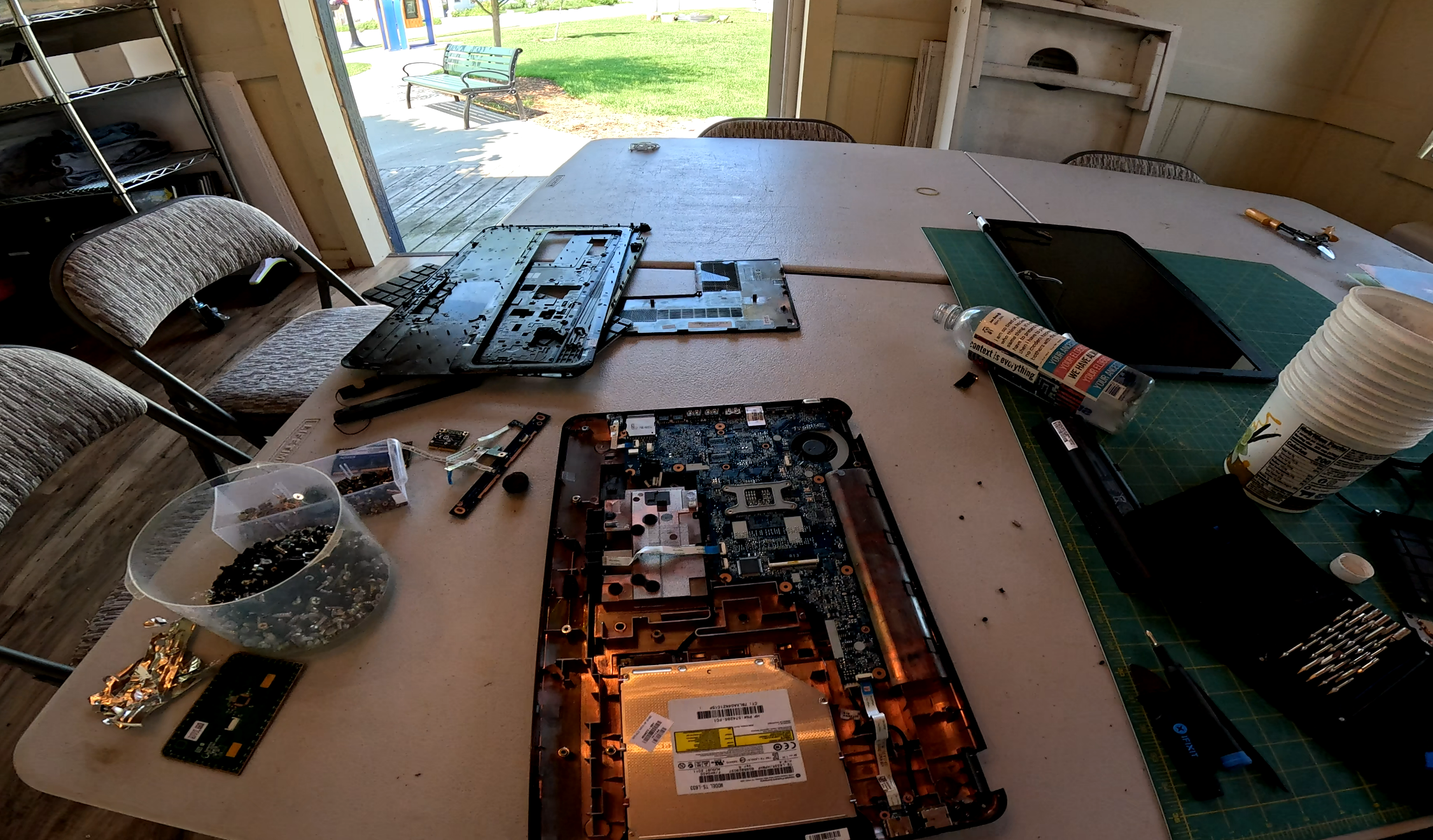 A mostly-disassembled laptop with its motherboard exposed, sitting on the big table in the workshop. Out the door of the workshop, the grass is green and the weather is sunny.