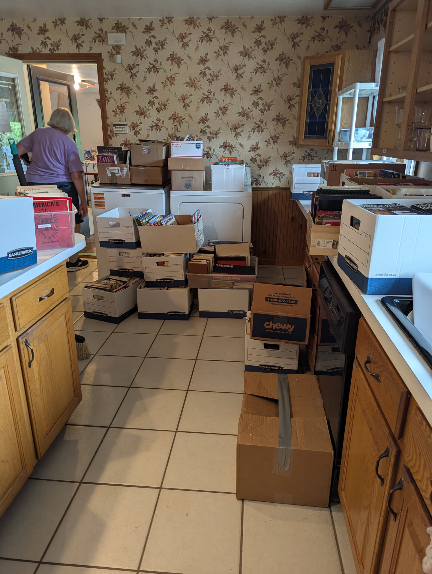 A kitchen with oak cabinets, white countertops, and a white tile floor. Every horizontal surface is covered in boxes of books.