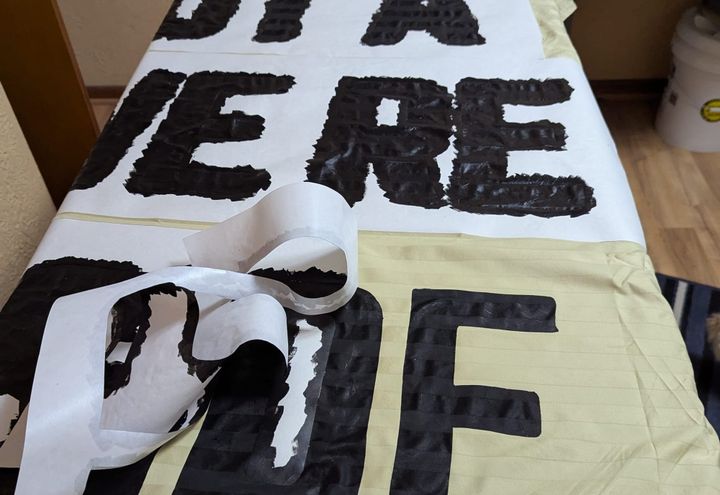 A banner made out of a striped satin sheet is being turned into a banner with a freezer paper stencil. The full text is not visible.