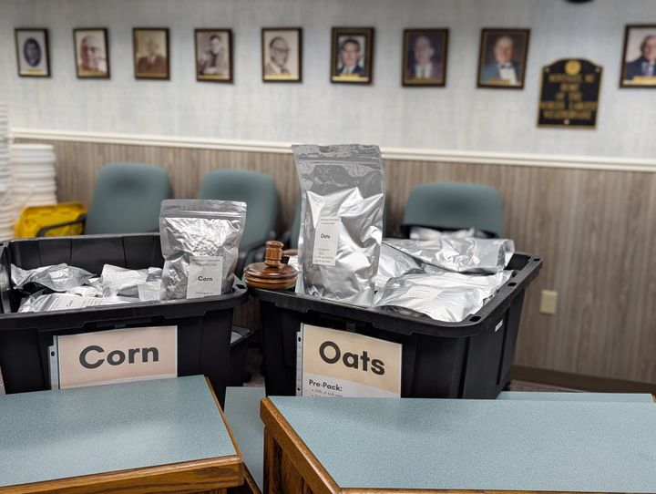 Two bins of mylar food packets are sitting on the city council desk. One says "Corn" and one says "Oats". The council gavel is positioned to make it look like a bag of oats is holding it.