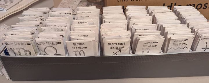 A long thin box made out of plastic from yard signs and covered in duct tape. It is divided into seven compartments, and each compartment holds packets of Astrology-themed tea