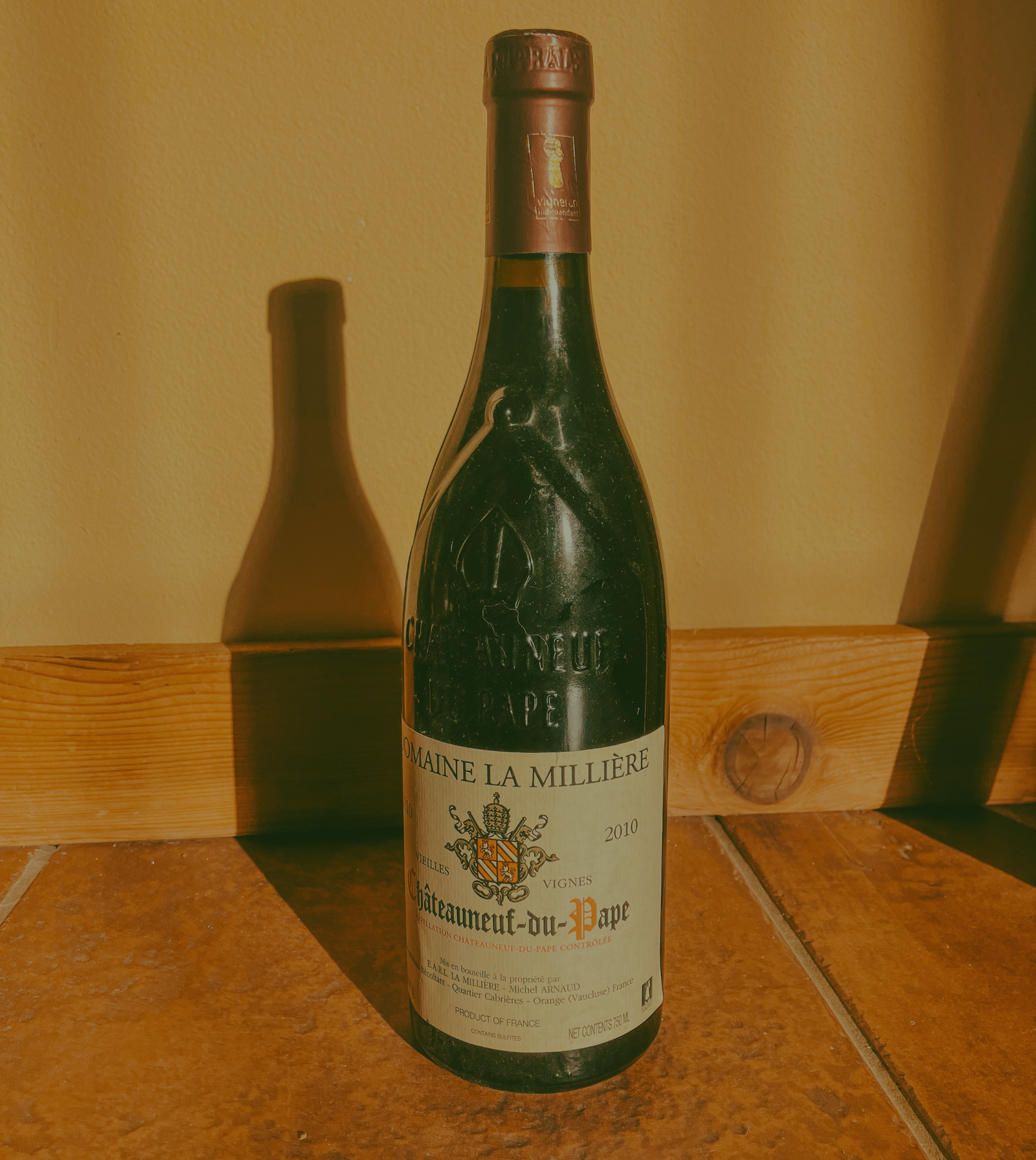 A bottle 2010 Domaine La Millière Châteauneuf-du-Pape Vieilles Vignes resting on tile floor, casting long shadow against the wall.