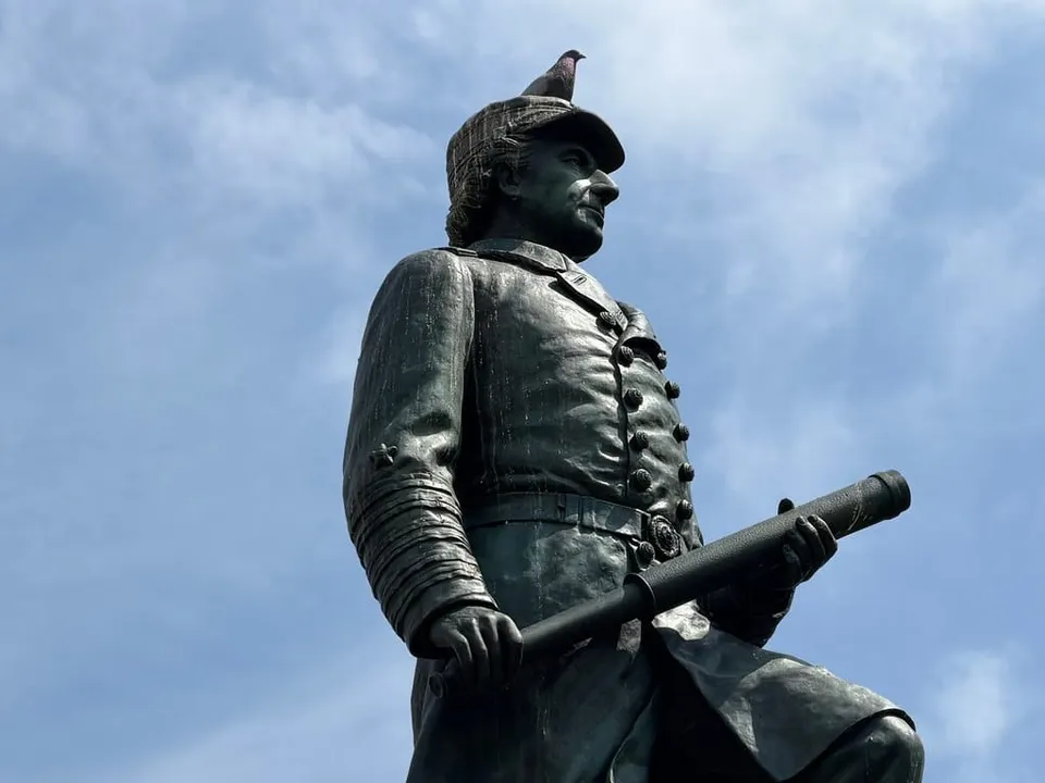 A statue with a pigeon sitting on its head. There is also blue sky behind the statue and pigeon. 