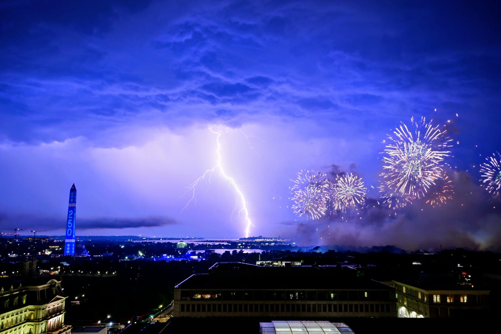 A lightning bolt near the Potomac River with fireworks to its right and the Washington Monument also visible.