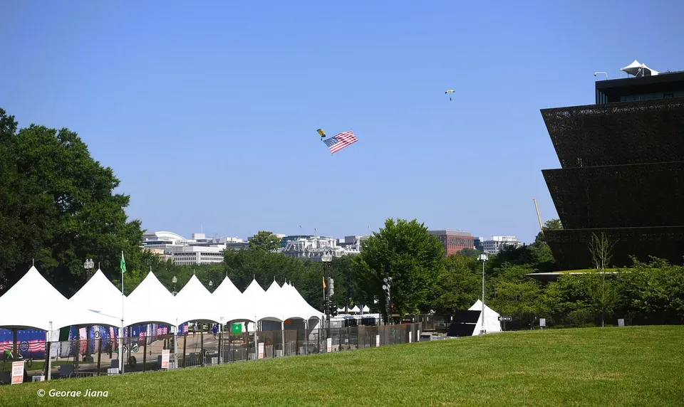 A parachute guy with an American flag over the National Mall on a blue sky day with setup for the Fourth. 
