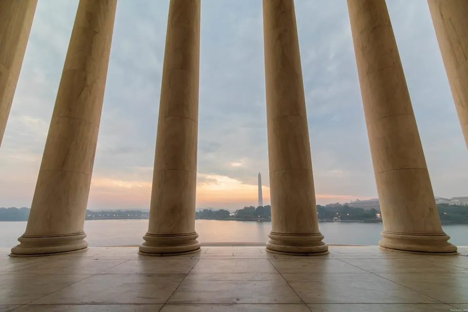 Looking through the columns of the Jefferson Memorial. It is cloudy. The Washington Monument is in the background. 