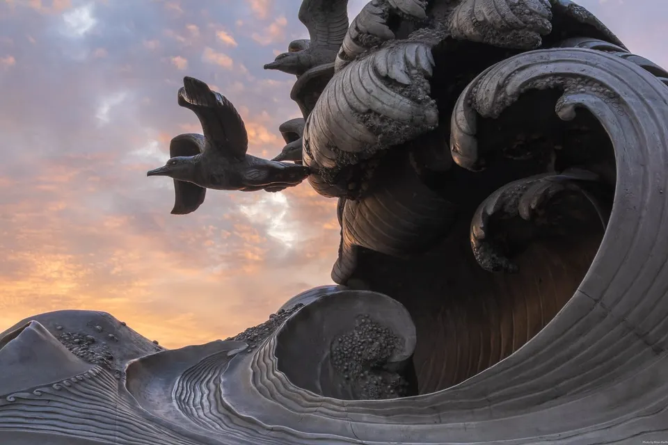 A statue with waves and birds and colorful morning clouds in the background.