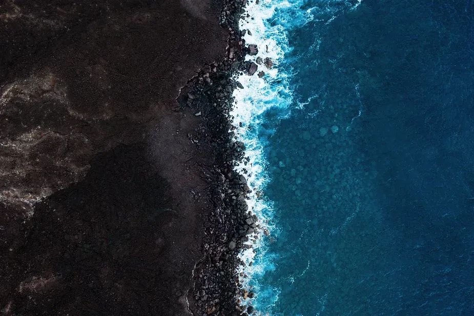 Color pic of aerial photo of a dark gray rocky shoreline on which brilliant blue water breaks into white froth.
