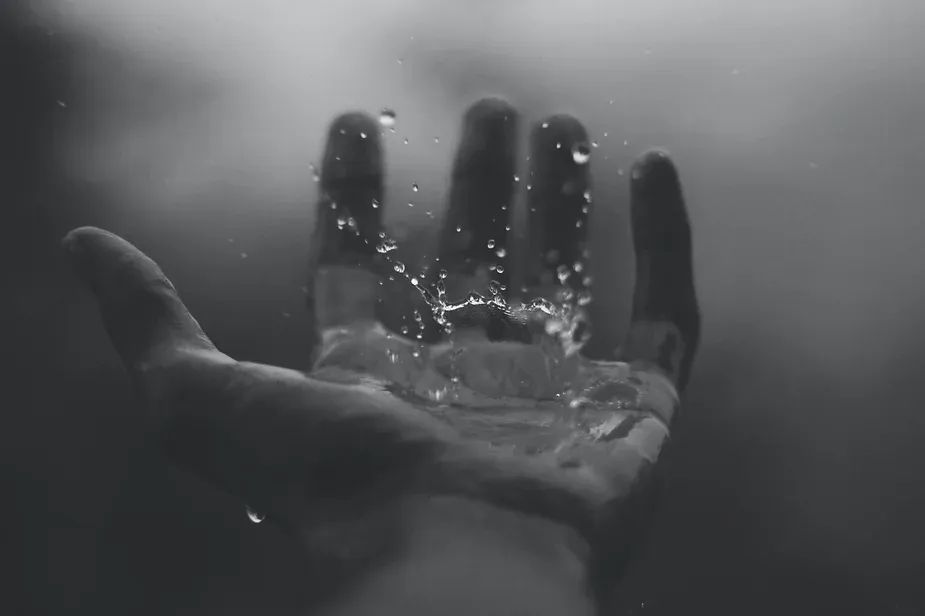 B/W photo of a masculine hand against a blurry background, with a water drop caught mid-splash in the palm