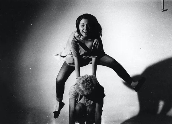 BW photo of two femme young adults playing leap-frog. The leaper, legs and clothes akimbo, has a wide grin